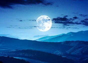Full moon rising over blue night landscape of rolling hills and mountains.