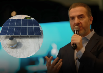 Speaker in a dark suit speaks into a microphone on stage; circular inset shows rooftop solar panels and a storage tank for solar energy.