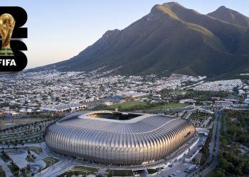Aerial view of a modern silver stadium beside a city and green hills, with a FIFA World Cup logo in the top-left corner.