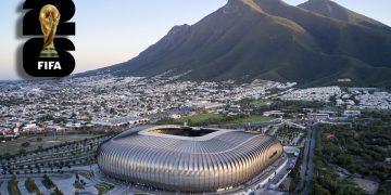 Aerial view of a modern silver stadium beside a city and green hills, with a FIFA World Cup logo in the top-left corner.