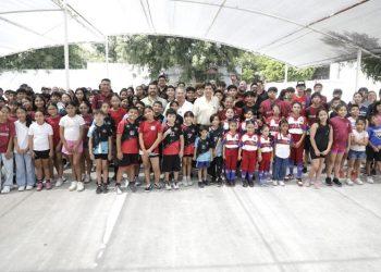 Large group of children and adults posing for a group photo under a translucent canopy outdoors; many are wearing sports uniforms, suggesting a school or club event.