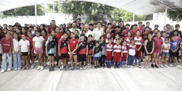 Large group of children and adults posing for a group photo under a translucent canopy outdoors; many are wearing sports uniforms, suggesting a school or club event.