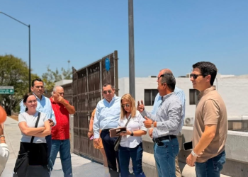Group of adults standing on a sunny rooftop, listening as a man gestures while speaking, others holding notebooks or bags.