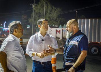 Three men stand at a nighttime construction site, conversing near orange traffic barrels and a large truck in the background.