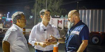 Three men stand at a nighttime construction site, conversing near orange traffic barrels and a large truck in the background.