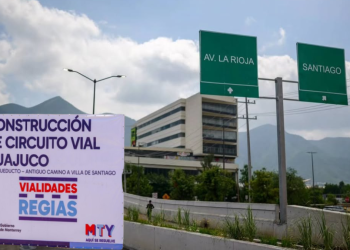 Banner announcing Huajuco road circuit construction with logos beside a highway, mountains in the background, and green directional signs reading AV. LA RIOJA and SANTIAGO.
