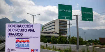 Banner announcing Huajuco road circuit construction with logos beside a highway, mountains in the background, and green directional signs reading AV. LA RIOJA and SANTIAGO.