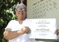 Smiling elderly woman wearing glasses and a white blouse holds a certificate outdoors, with green plants and a mural in the background.