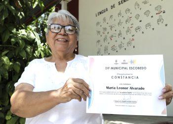 Smiling elderly woman wearing glasses and a white blouse holds a certificate outdoors, with green plants and a mural in the background.