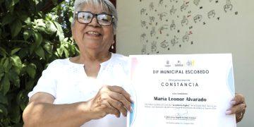 Smiling elderly woman wearing glasses and a white blouse holds a certificate outdoors, with green plants and a mural in the background.